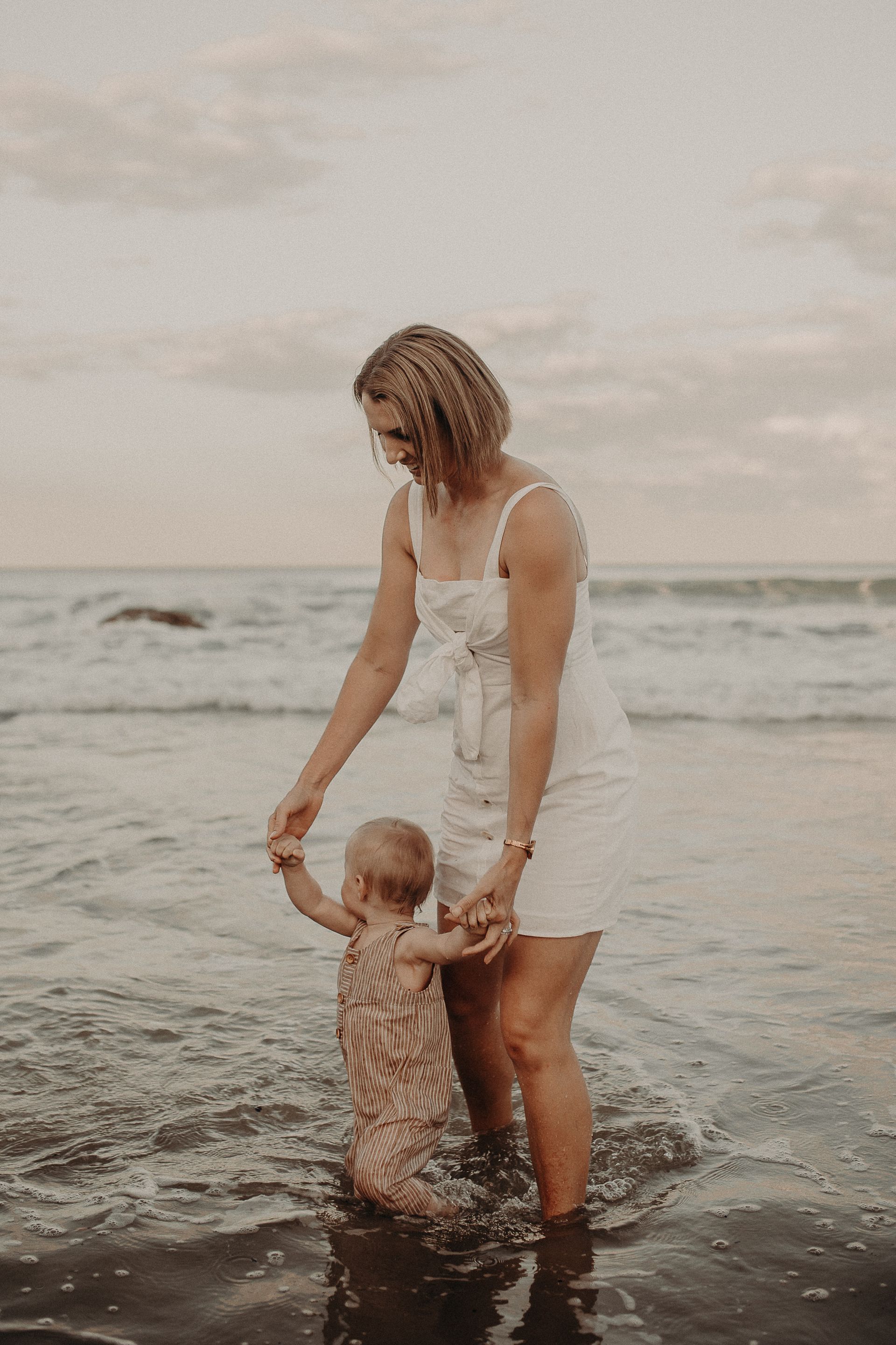Woman and baby in the ocean holding hands, smiling. Sky is overcast, waves are gentle.