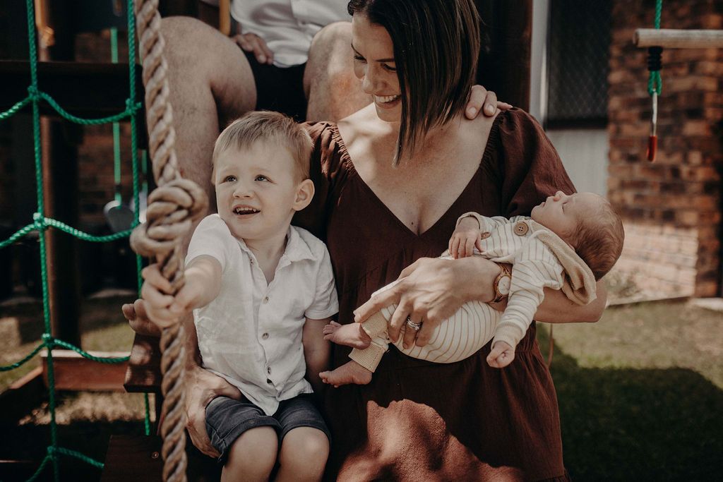 Woman holds a baby, with a boy beside her. They smile in a backyard, near a playground.