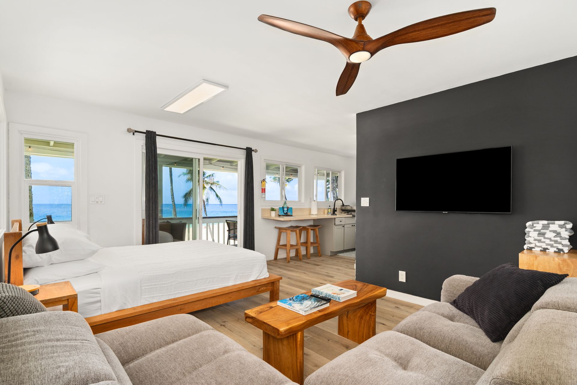 Cozy studio apartment with ocean view. Features: bed, sofa, TV on dark wall, ceiling fan, and small kitchen.