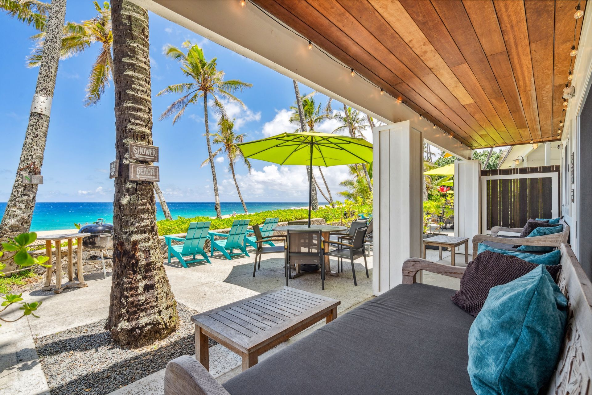 Covered patio overlooking the ocean with seating, tables, and palm trees on a sunny day.