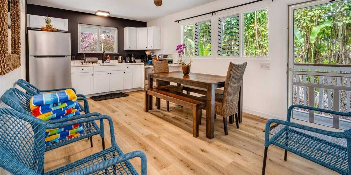 Cozy interior with blue wicker furniture, a wooden dining set, and a kitchen, with a view of lush greenery.