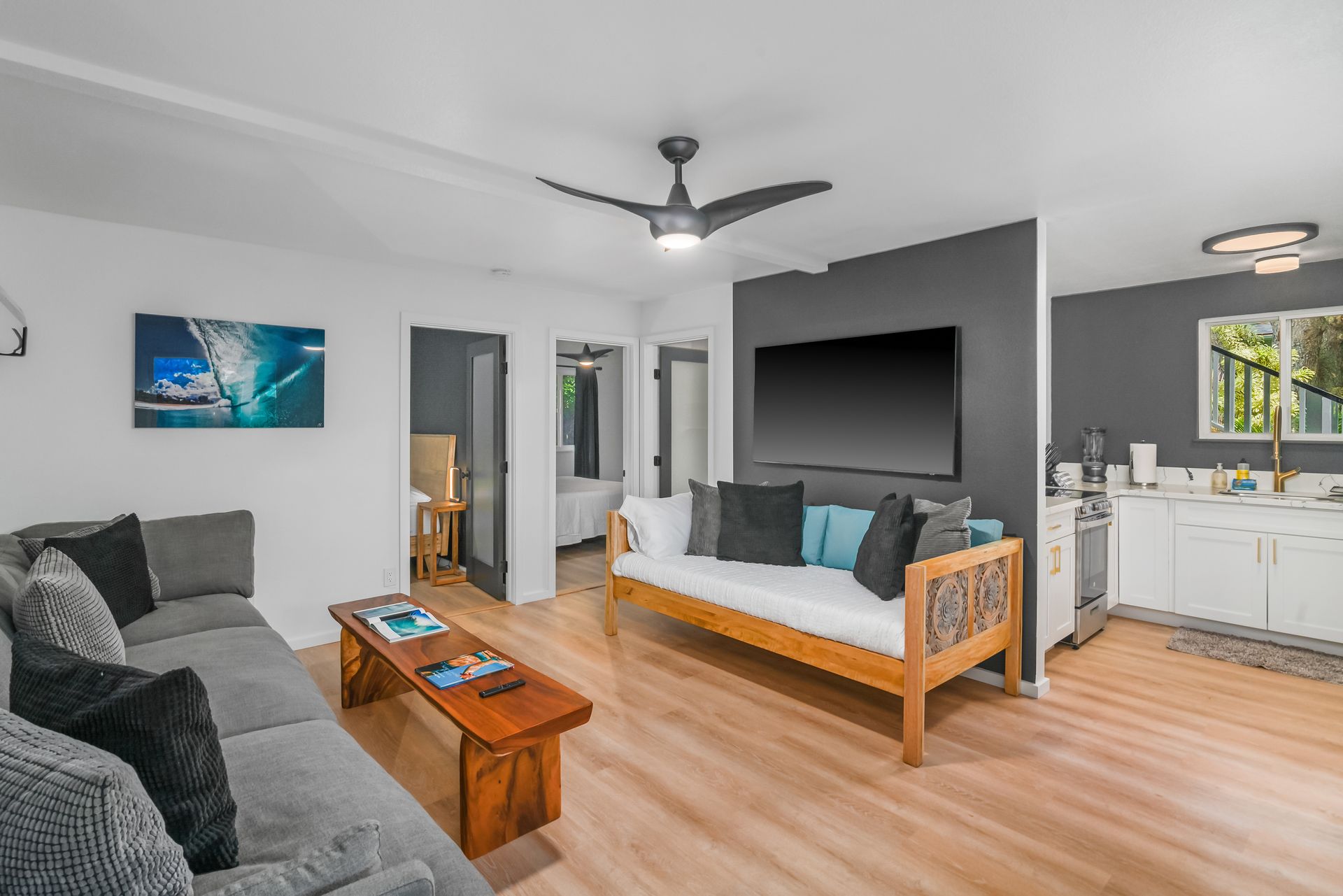 Living room with gray sofa, wooden daybed, TV, and kitchen visible.