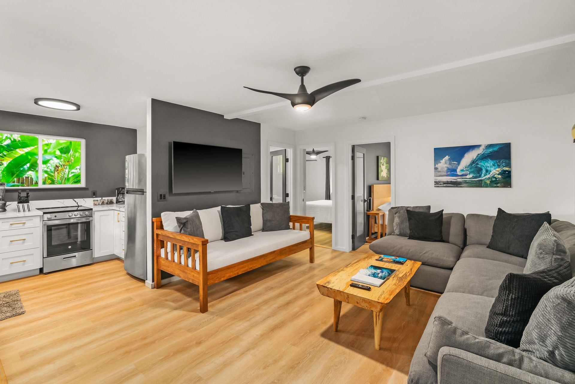 Open-plan living room with a kitchen, gray sectional sofa, wooden futon, TV, and light wood floors.