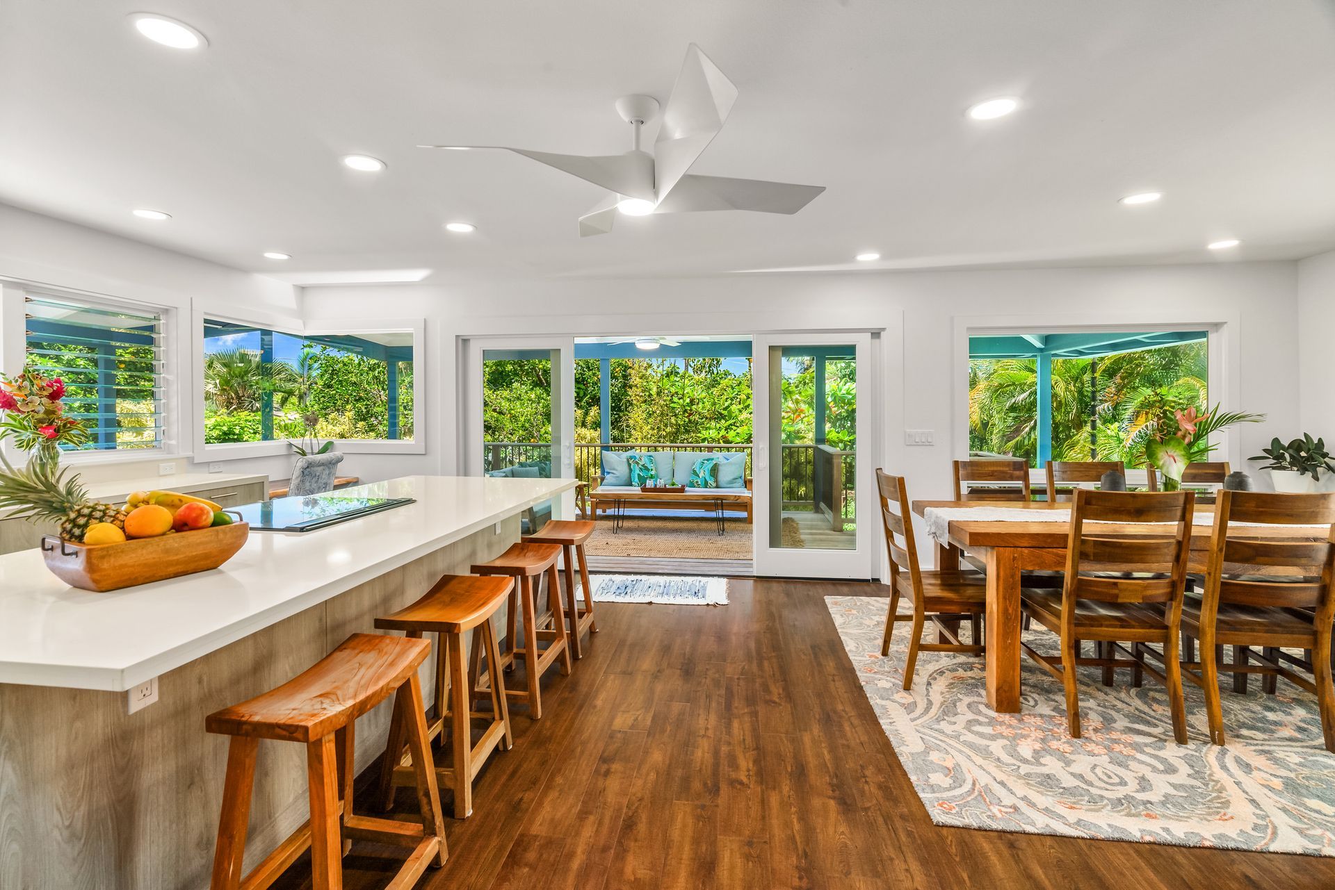 Bright open kitchen and dining area with a view of greenery. Wooden floor, island with bar stools, and a dining table.