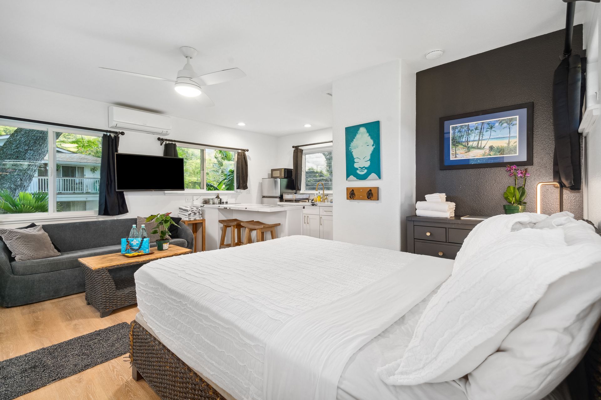 Bed in foreground, living area with sofa & TV, kitchen in background, white walls, wood floors.