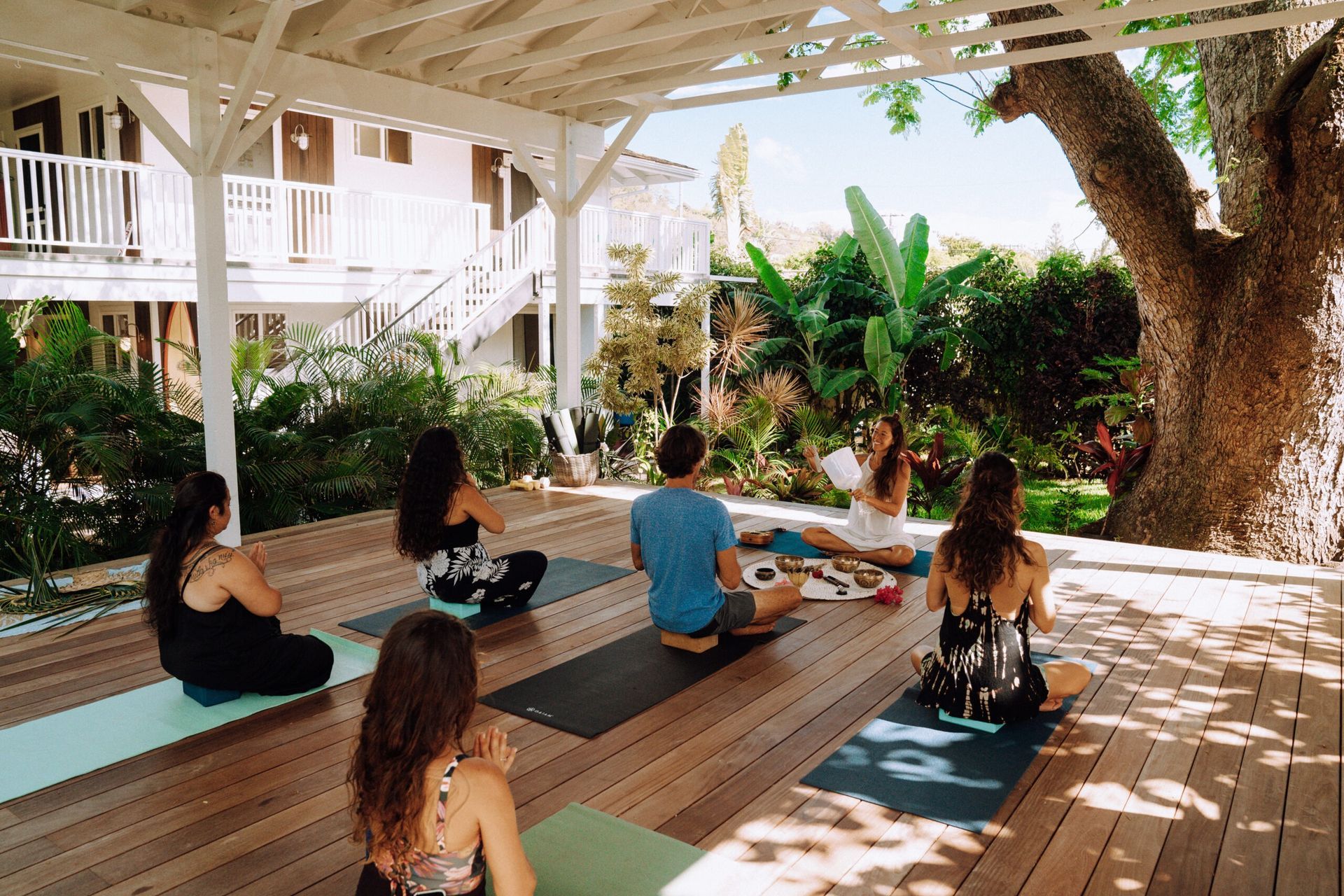 Yoga class outdoors on a wooden deck, instructor and students seated in meditation.