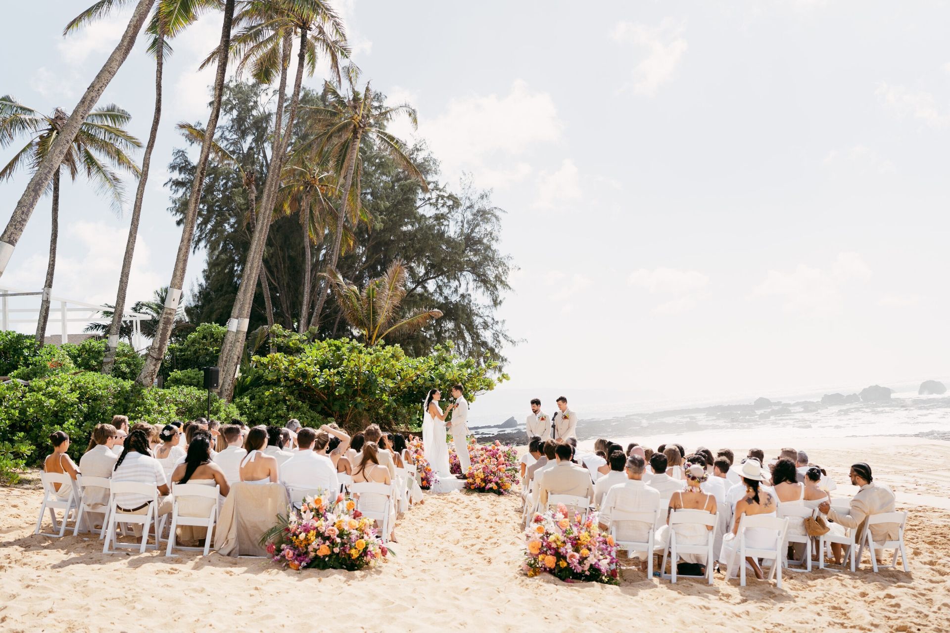 Wedding ceremony on a sandy beach with guests in white, palm trees, and colorful floral arrangements.