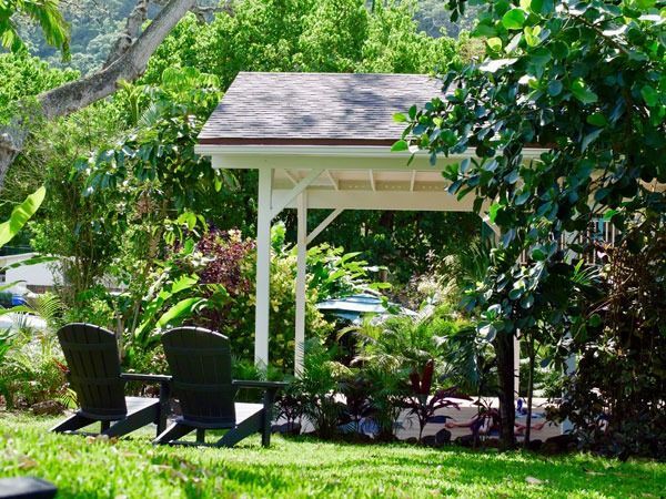 Two black Adirondack chairs under a white-framed shelter in a lush, green garden.