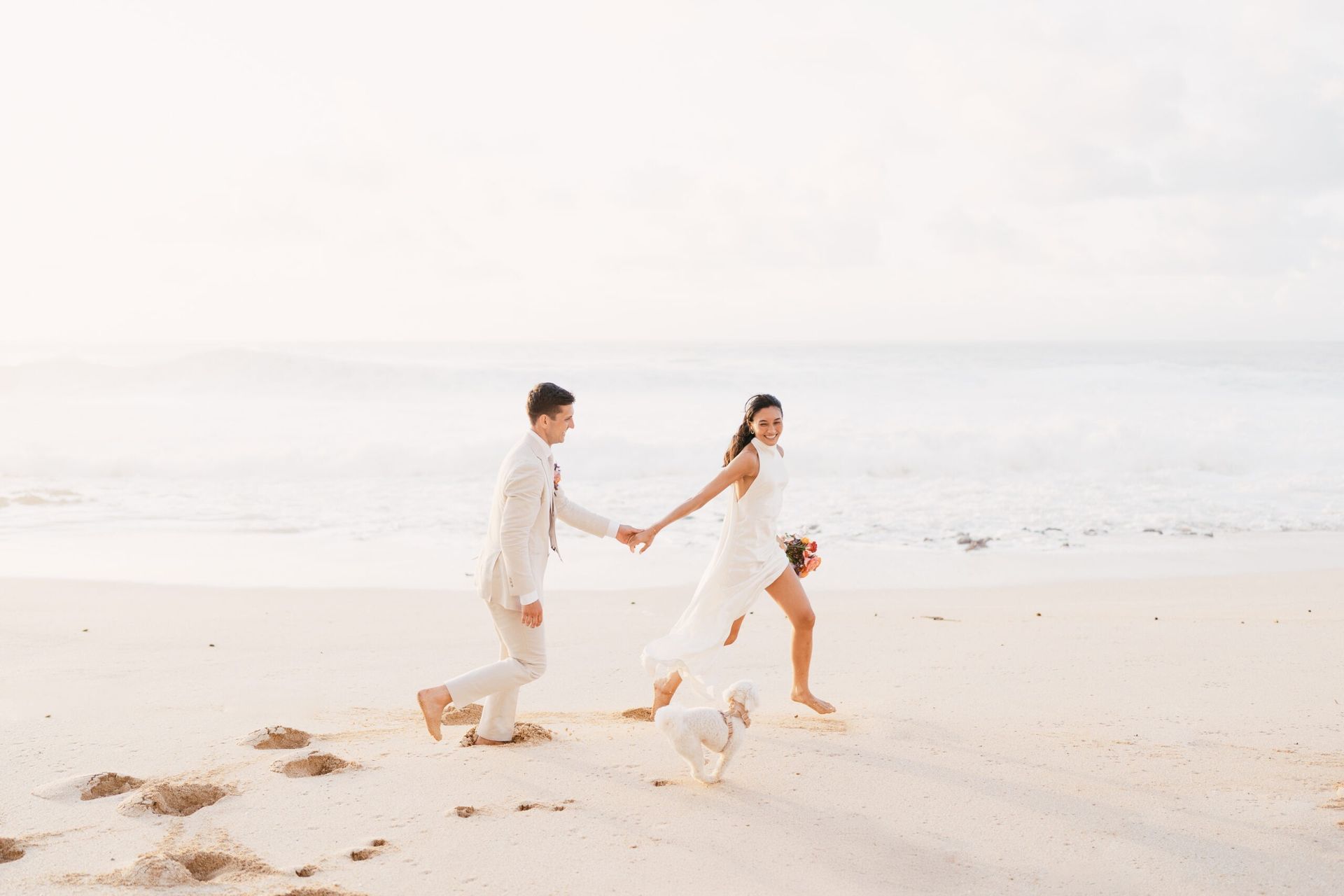 Bride and groom holding hands, running on a beach at sunset, with a small white dog following.