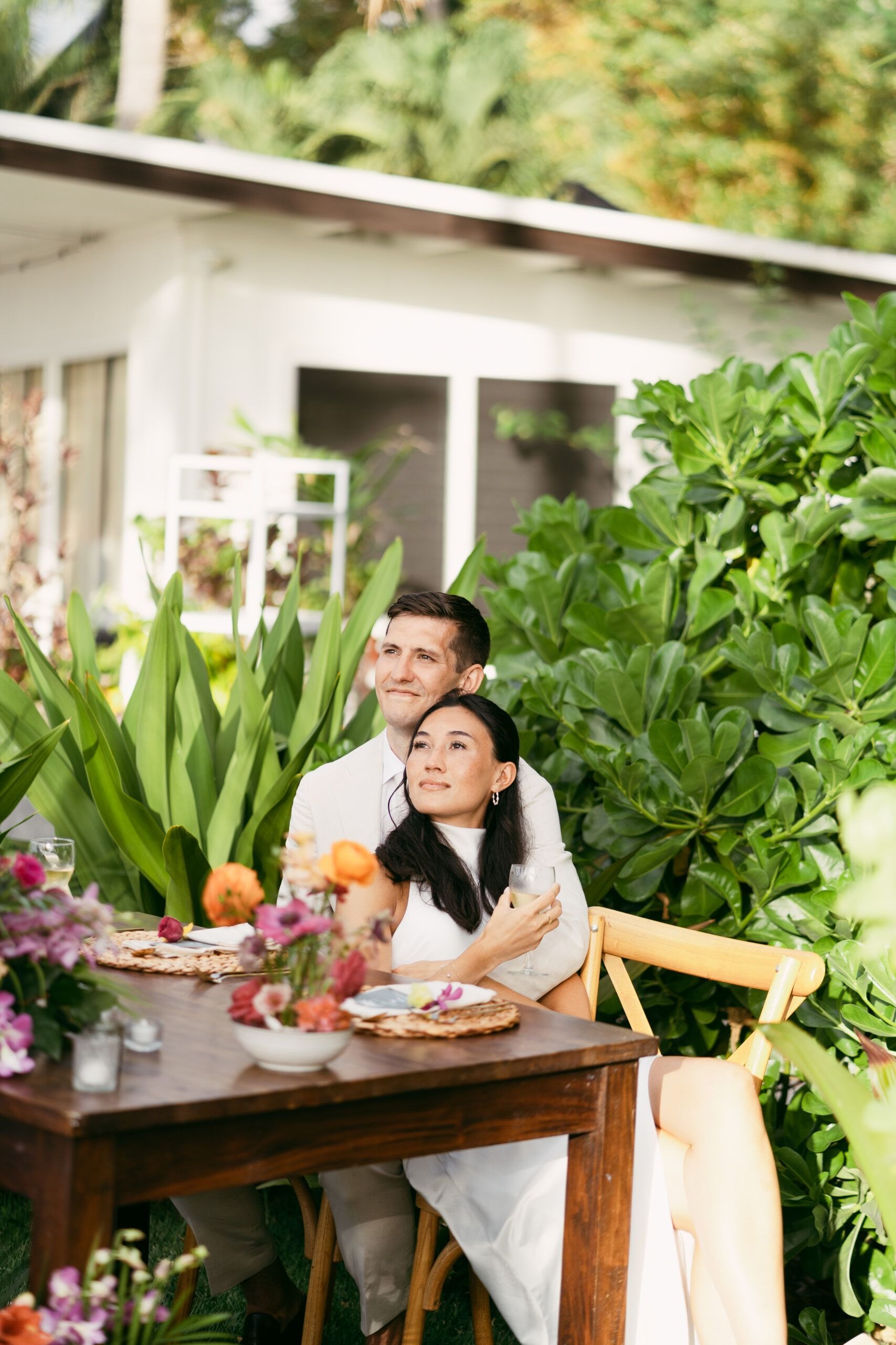 Couple embraces at outdoor table with flowers, looking upwards, near a white building.
