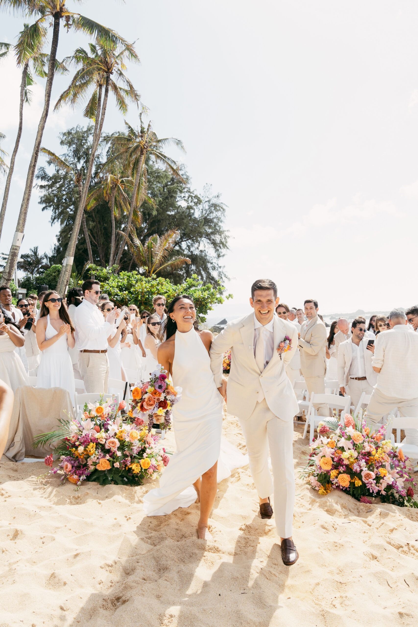 Newlyweds run down a beach aisle, smiling, toward guests; colorful flowers, palm trees.