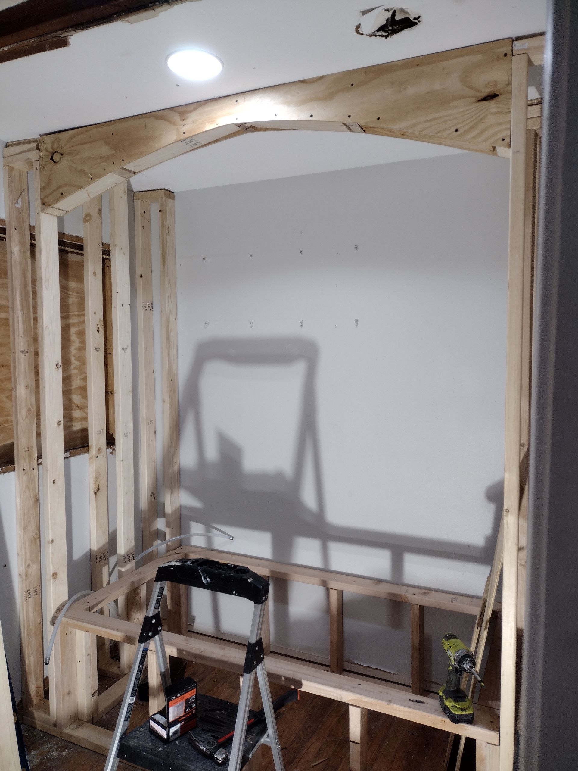 Interior room under construction, with wooden frame, archway, and small bench. A stepladder is present.
