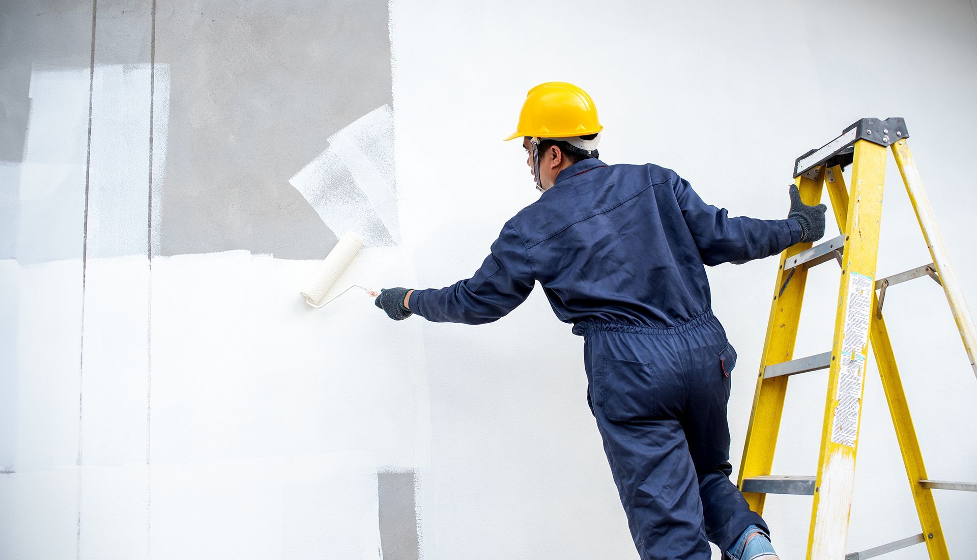 Person in blue coveralls and yellow hard hat painting a wall white with a roller from a yellow ladder.