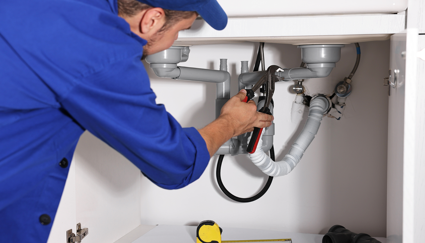 Plumber in blue uniform working under a sink, using pliers on pipes.