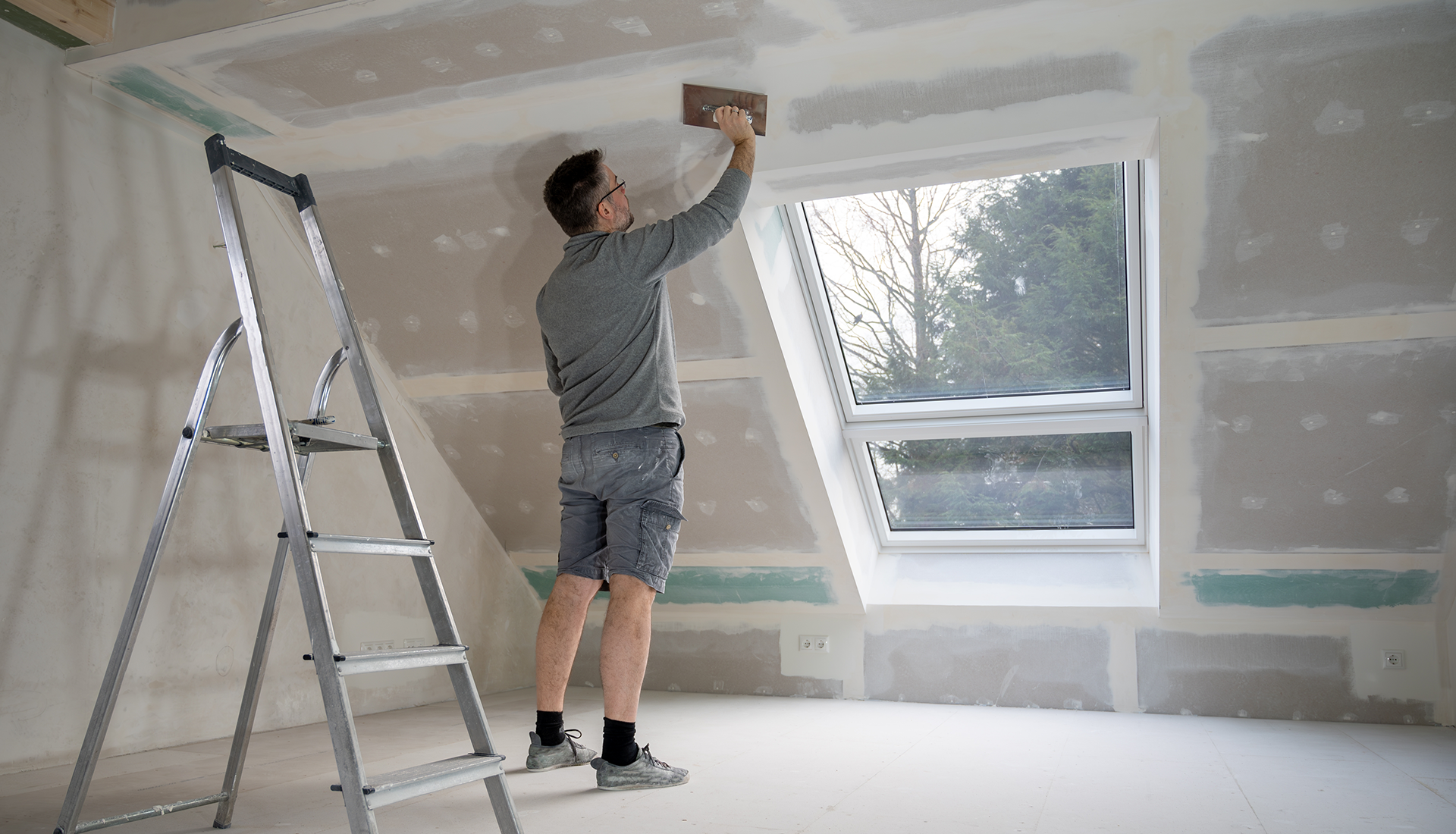 Person painting a room ceiling beside a ladder and large window