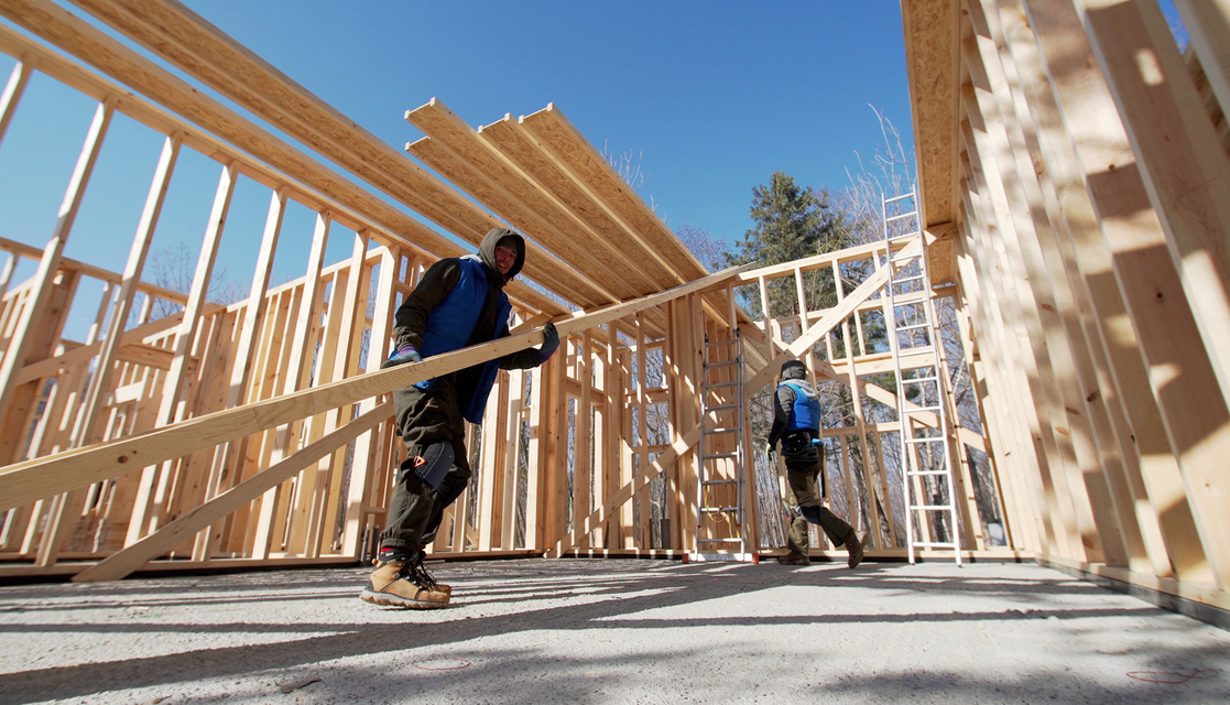 Two people building a wooden frame structure outdoors, carrying lumber. Blue sky overhead.
