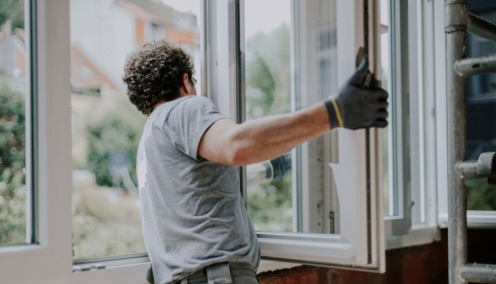 A person installing a white window in a building, wearing work gloves and a gray shirt.