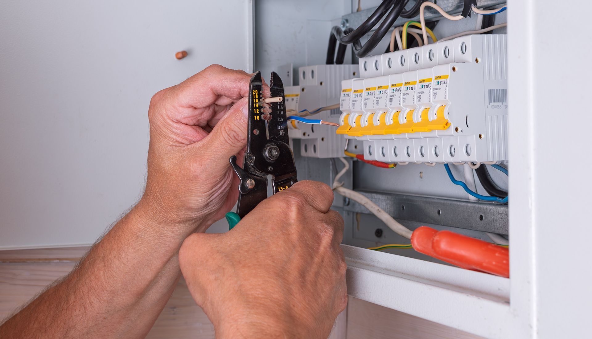 Hands using wire strippers on electrical wiring inside a circuit breaker box.