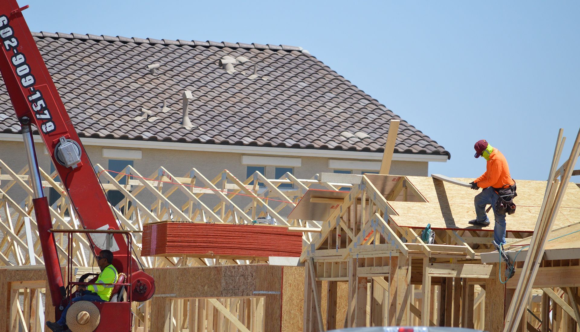 Construction workers building a house with a crane lifting lumber on a sunny day.