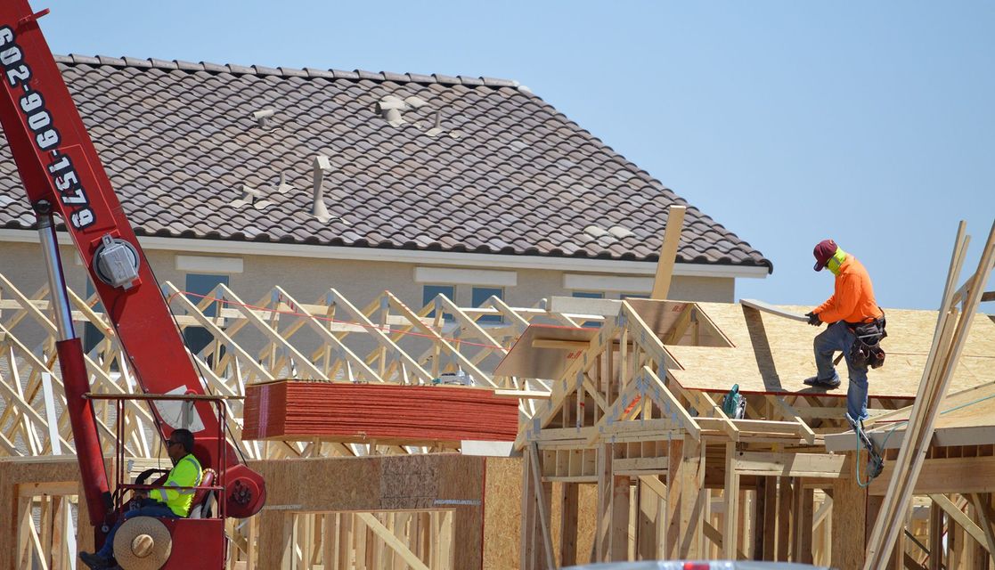 Construction workers building a house with a crane lifting lumber on a sunny day.