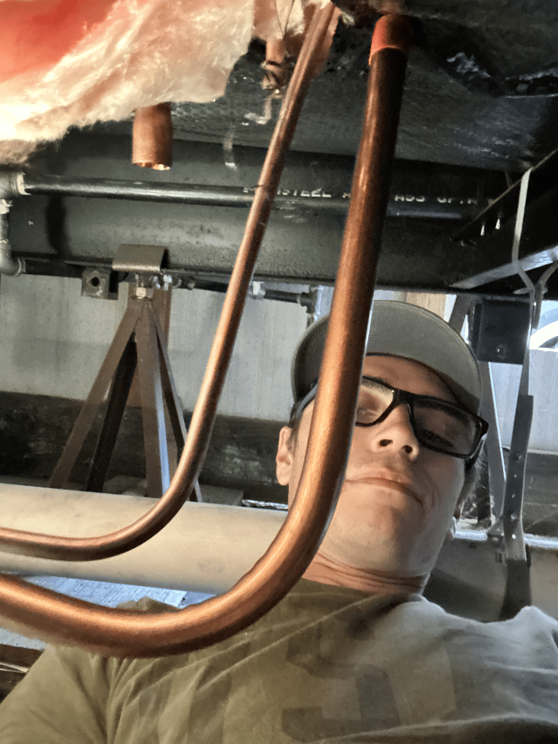 A man wearing glasses and a hat is working under a pipe