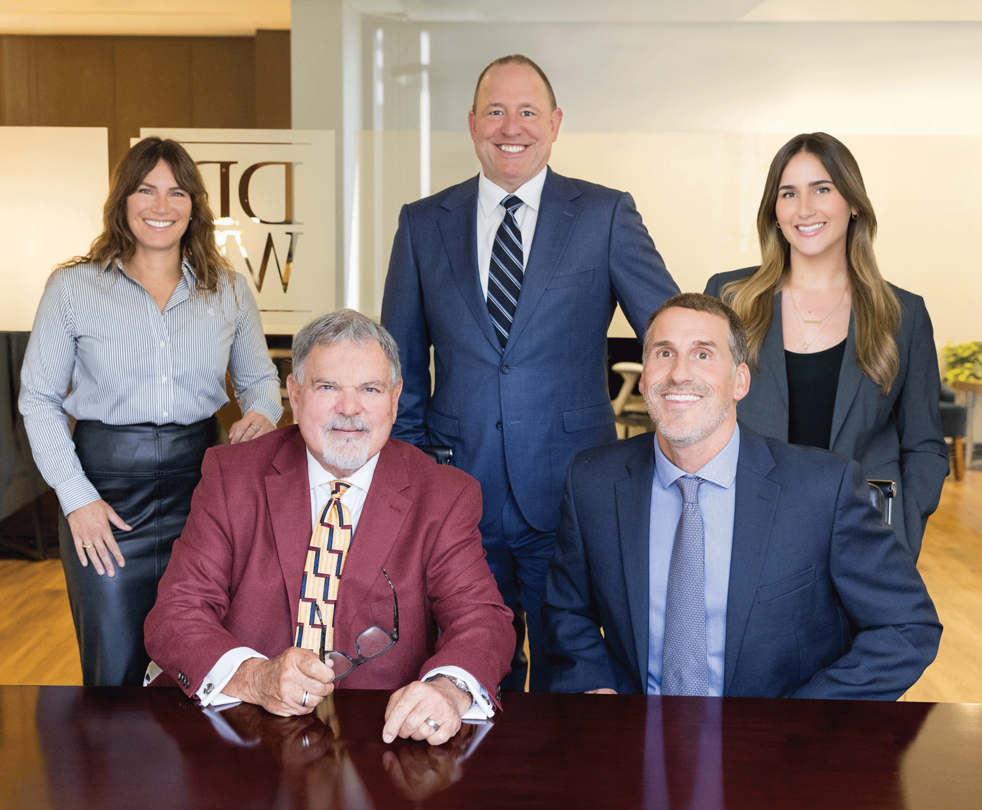 Five people in suits and professional attire posing at a dark wood table in an office setting.