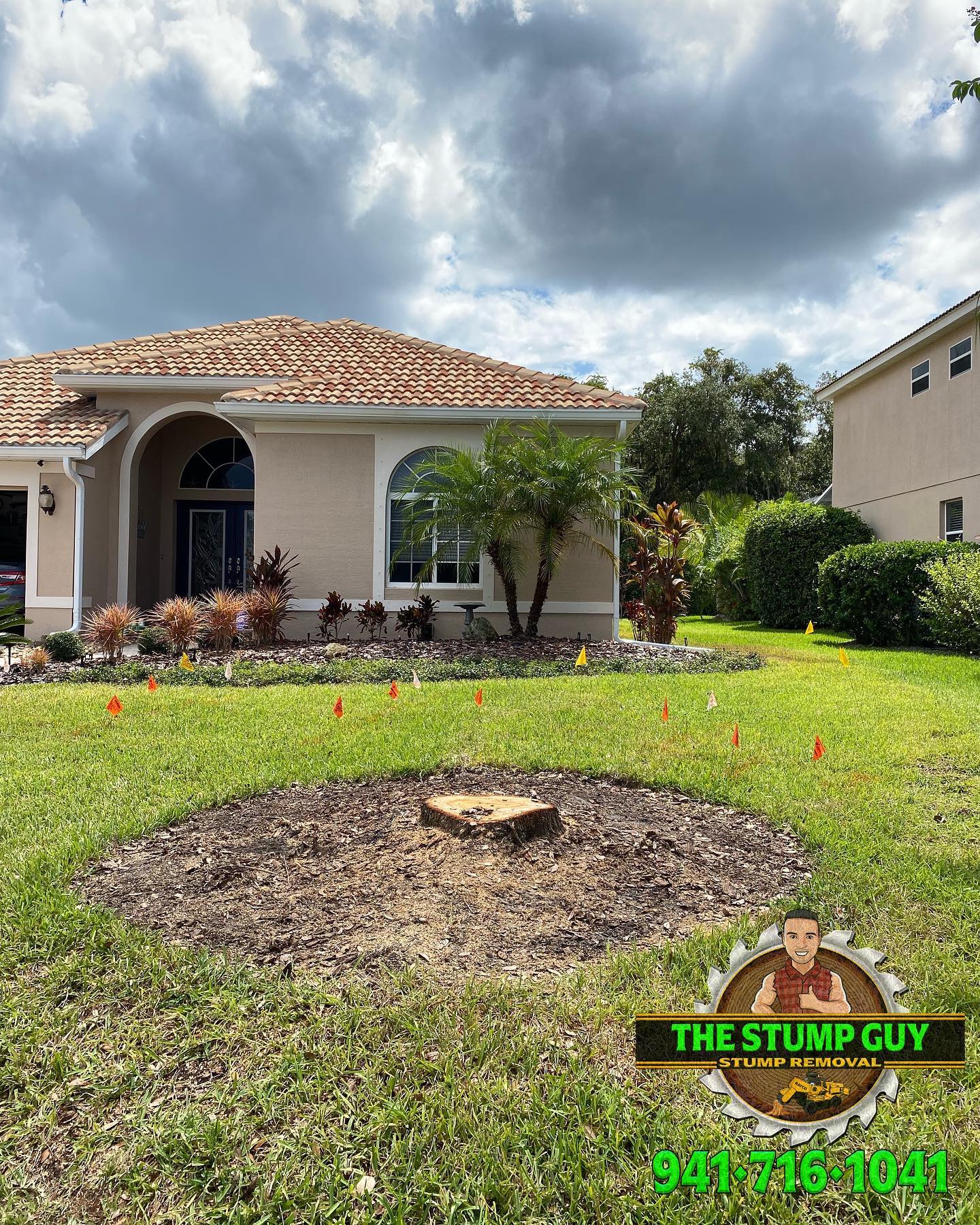 Stump removed from yard in front of beige house, surrounded by wood chips and orange flags. Green lawn, cloudy sky.