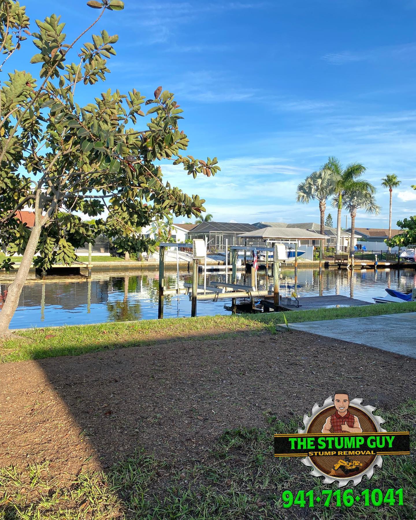Waterfront scene with a boat dock. Grass in the foreground, with a tree. Clear blue sky.