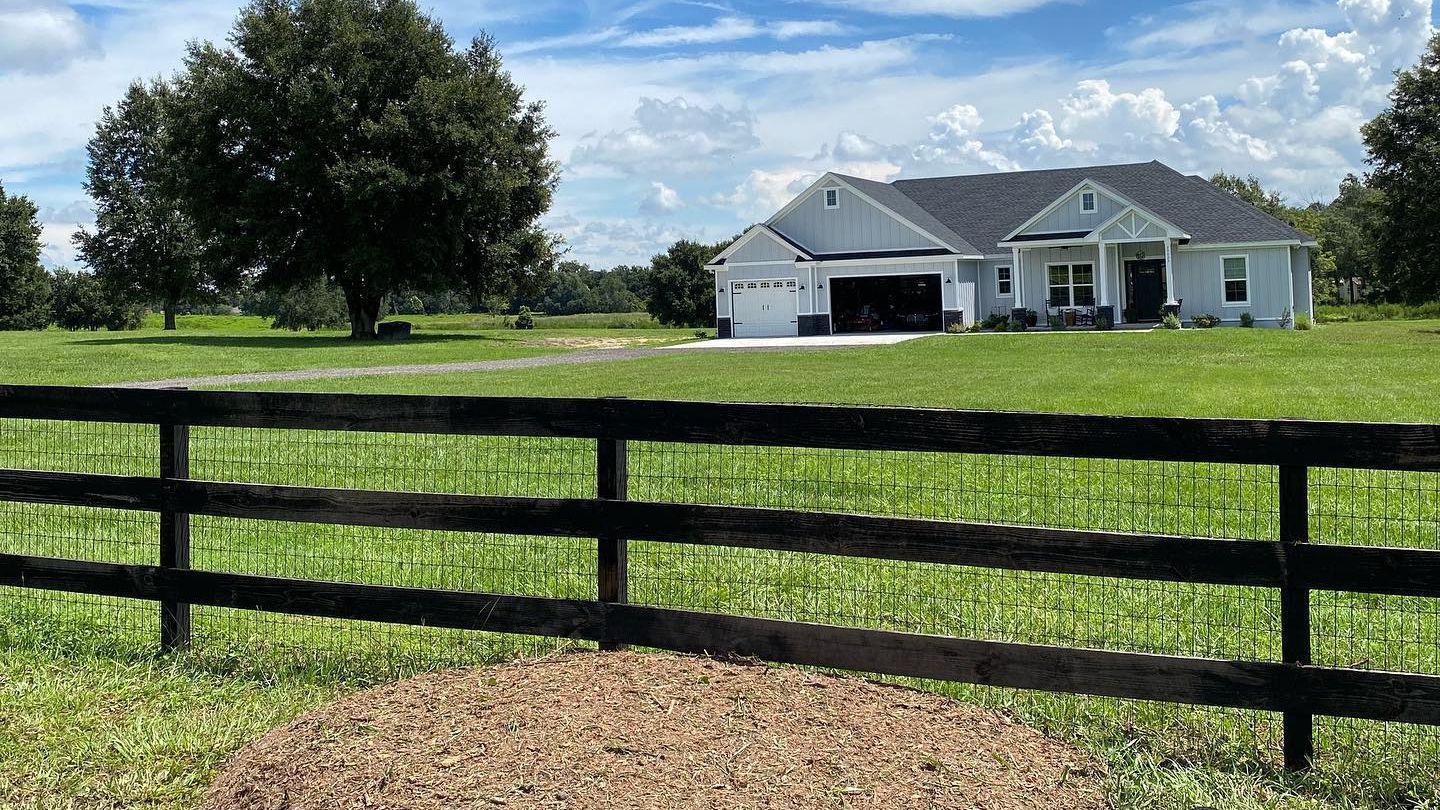 A black fence in front of a green lawn and white house under a blue sky with puffy clouds.