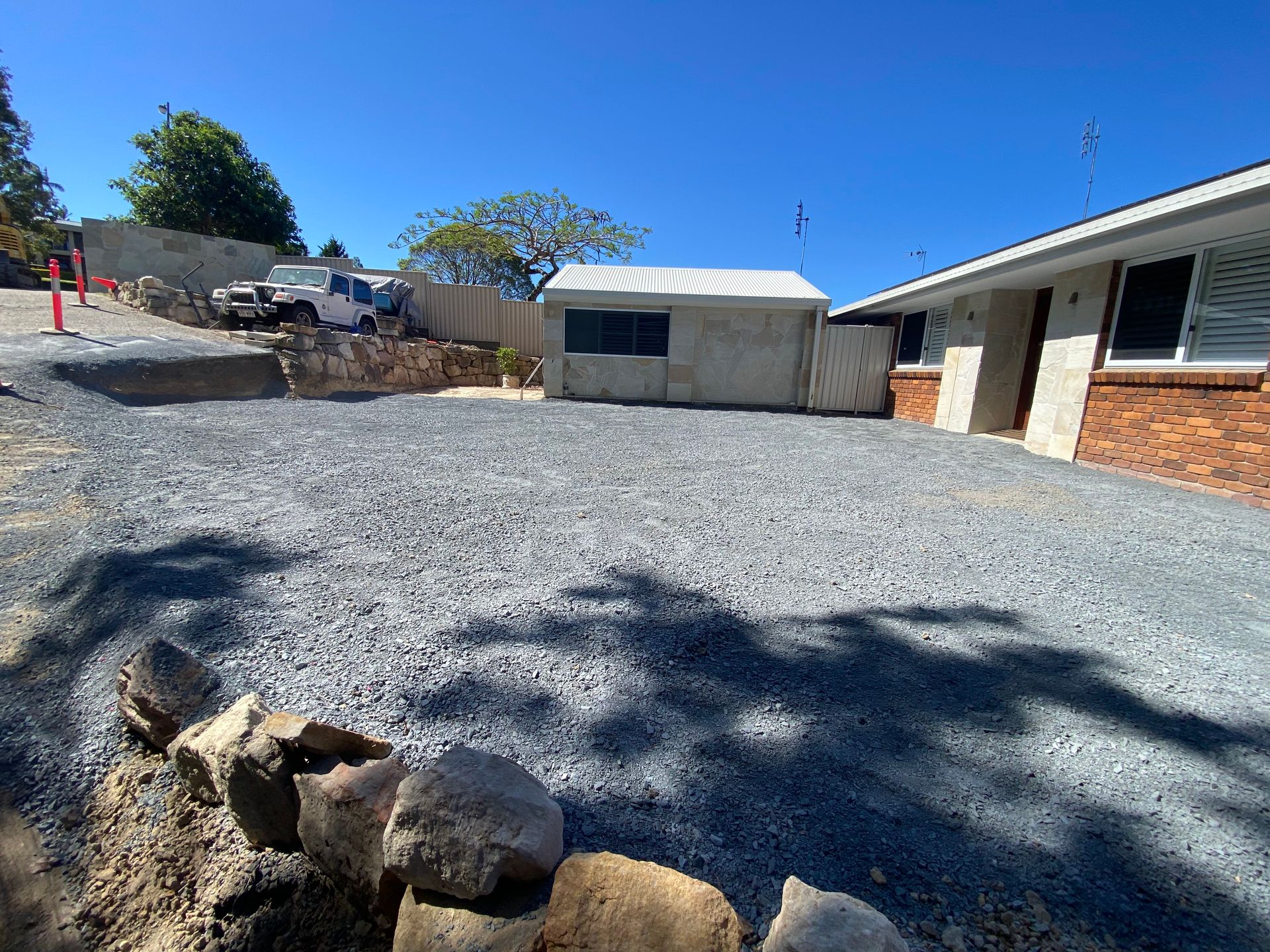 Gravel driveway in front of a house, small shed, and retaining wall made of rocks; blue sky.