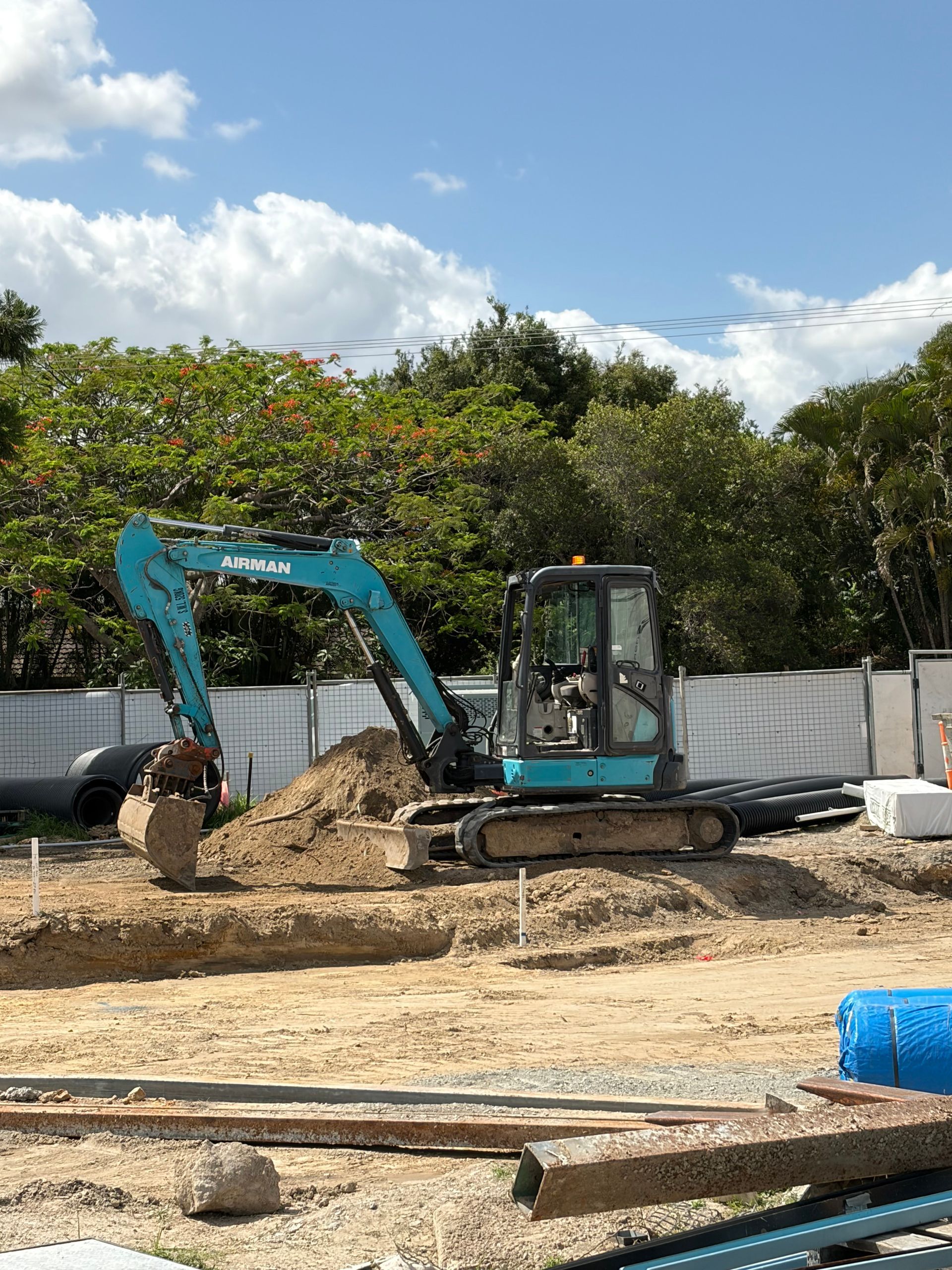 Blue excavator digging in a construction site with dirt and concrete blocks under a blue sky.