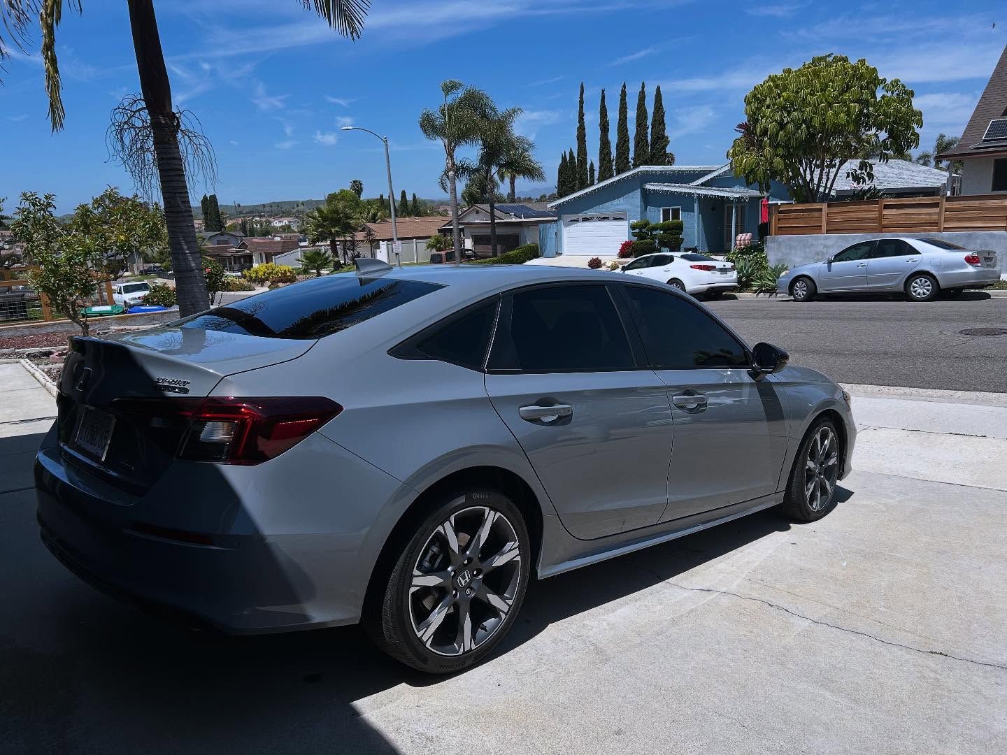 Gray sedan parked in a driveway on a sunny day. Houses and trees in the background.