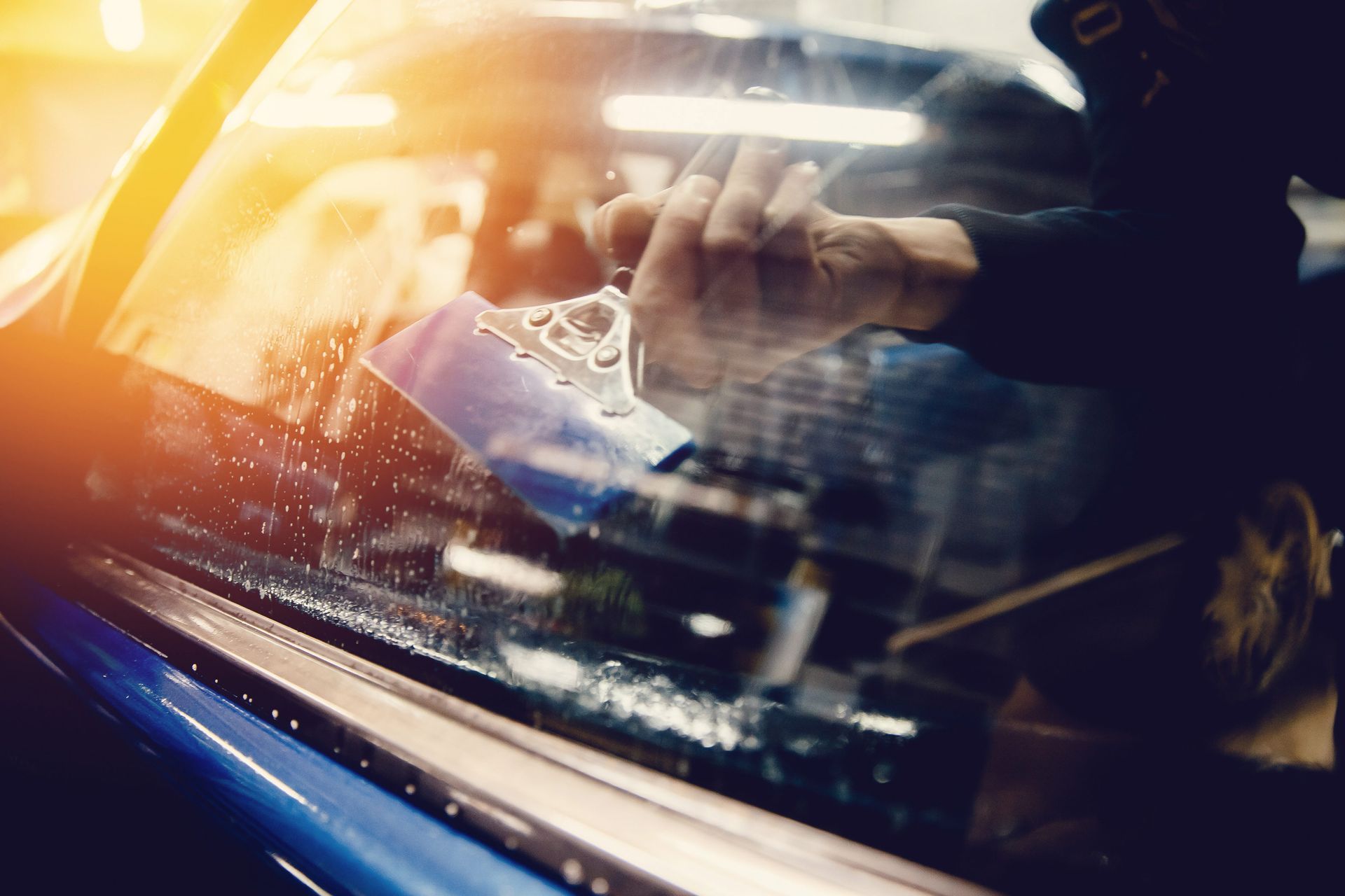 Person applying tint film to a car window. Blue car, hands using a tool, sunlight.