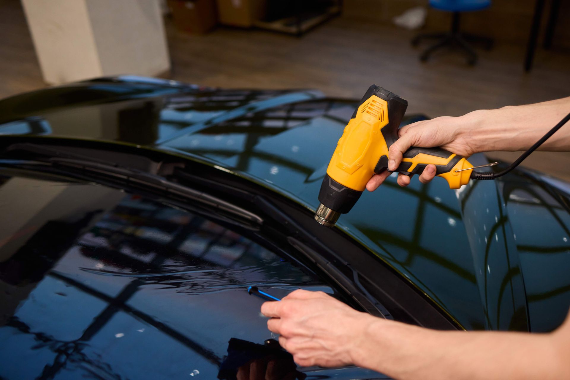 Person applying window tint to a car with a heat gun in a workshop.