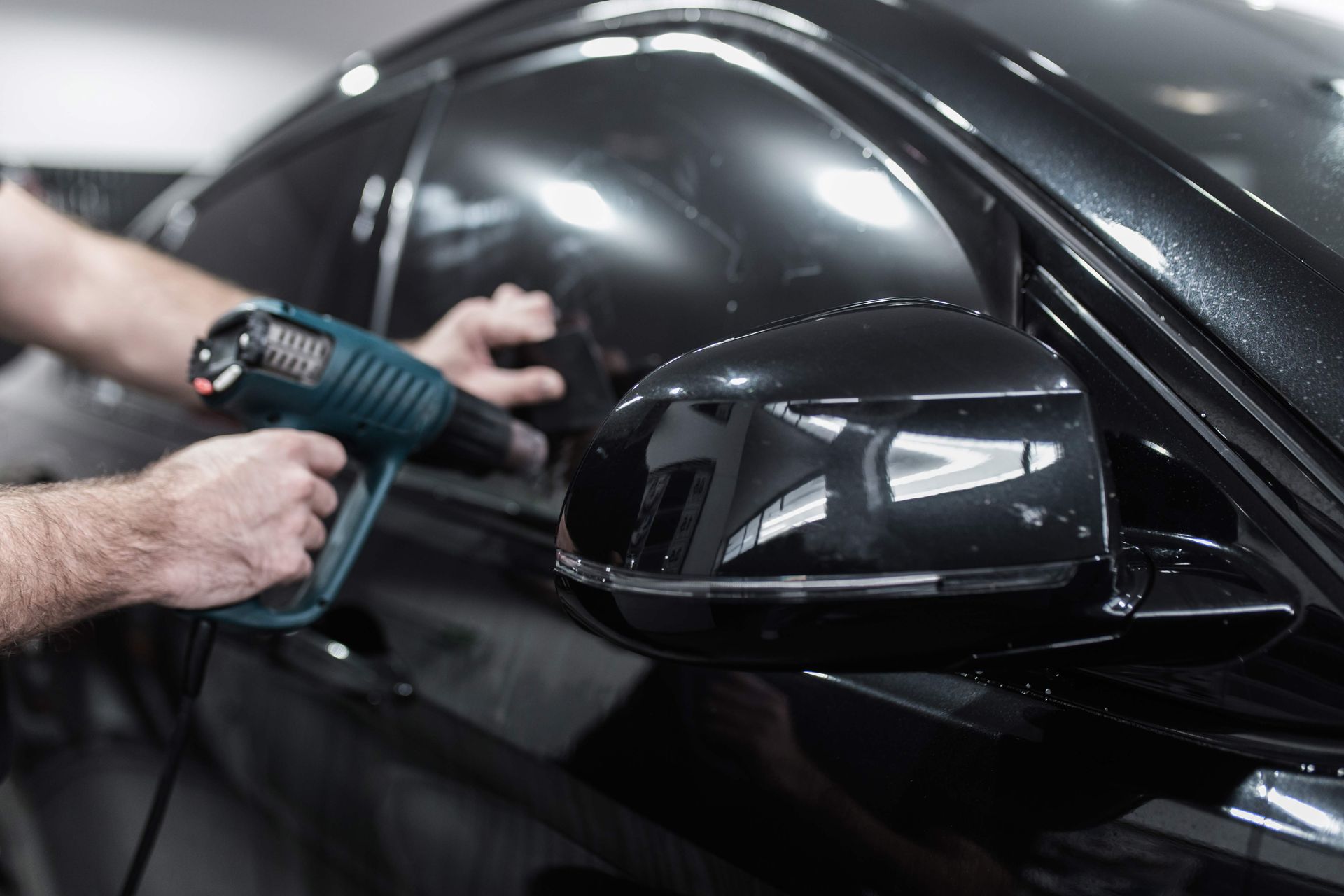 Person applying window tint to a black car with a heat gun, in a garage.