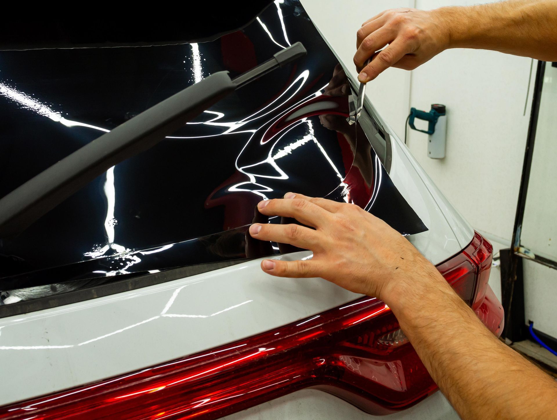 Person applying window tint to a car's rear window. Hands use tools to smooth film on white car.
