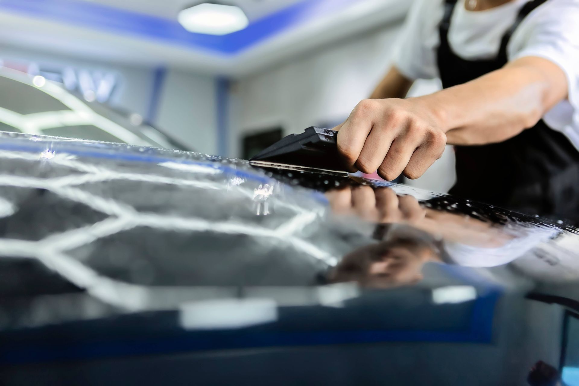 Person applying tint film to a car windshield with a squeegee in a brightly lit workshop.