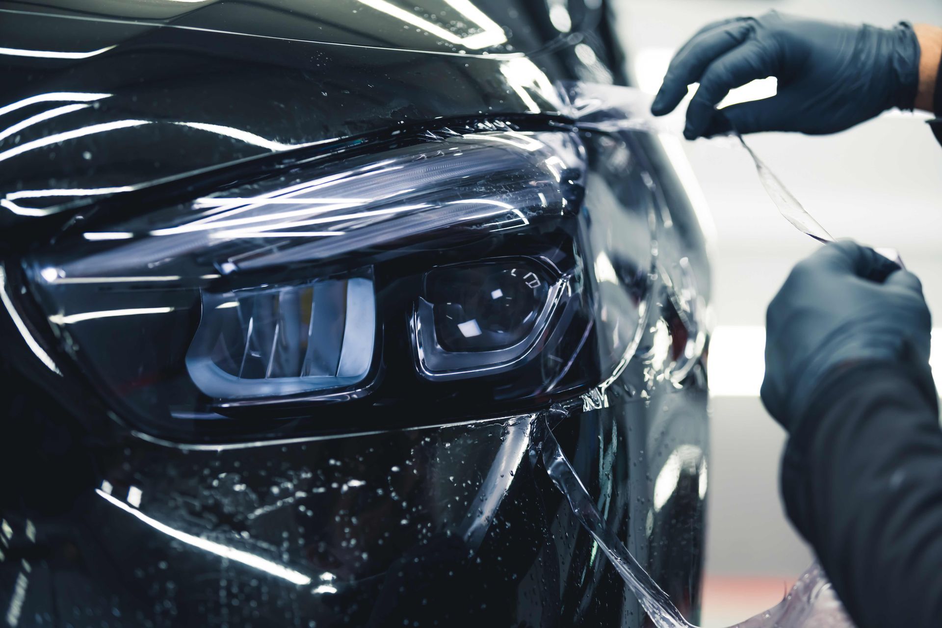 A person wearing black gloves applying protective film to a black car's headlight.