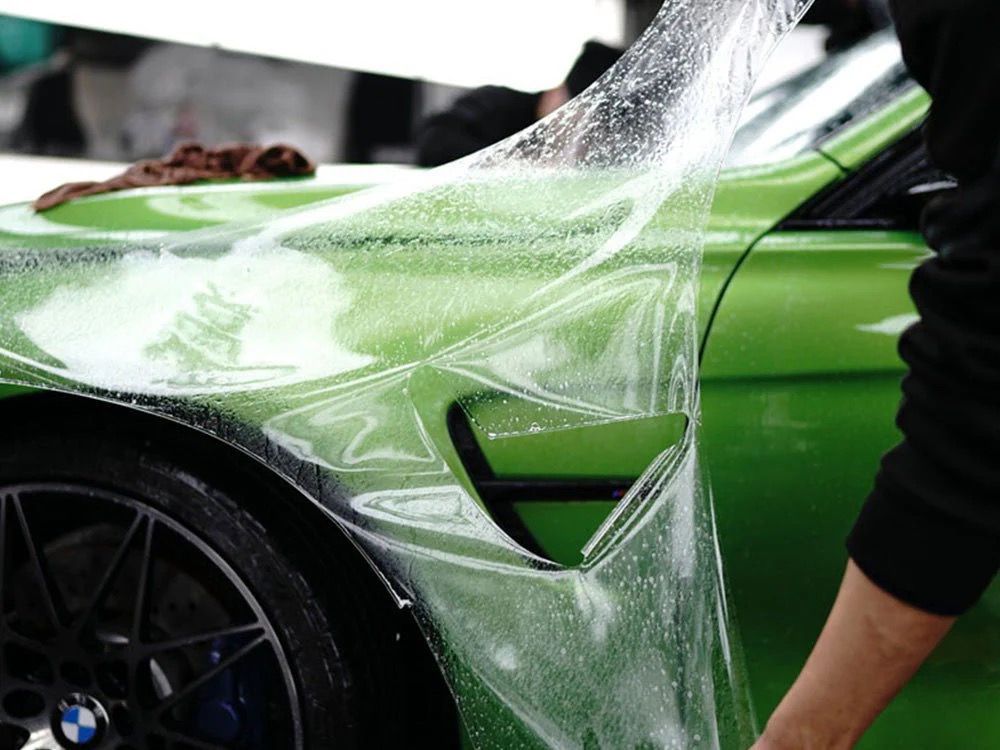 Hands in blue gloves applying ceramic coating to a black car with a sponge and bottle.