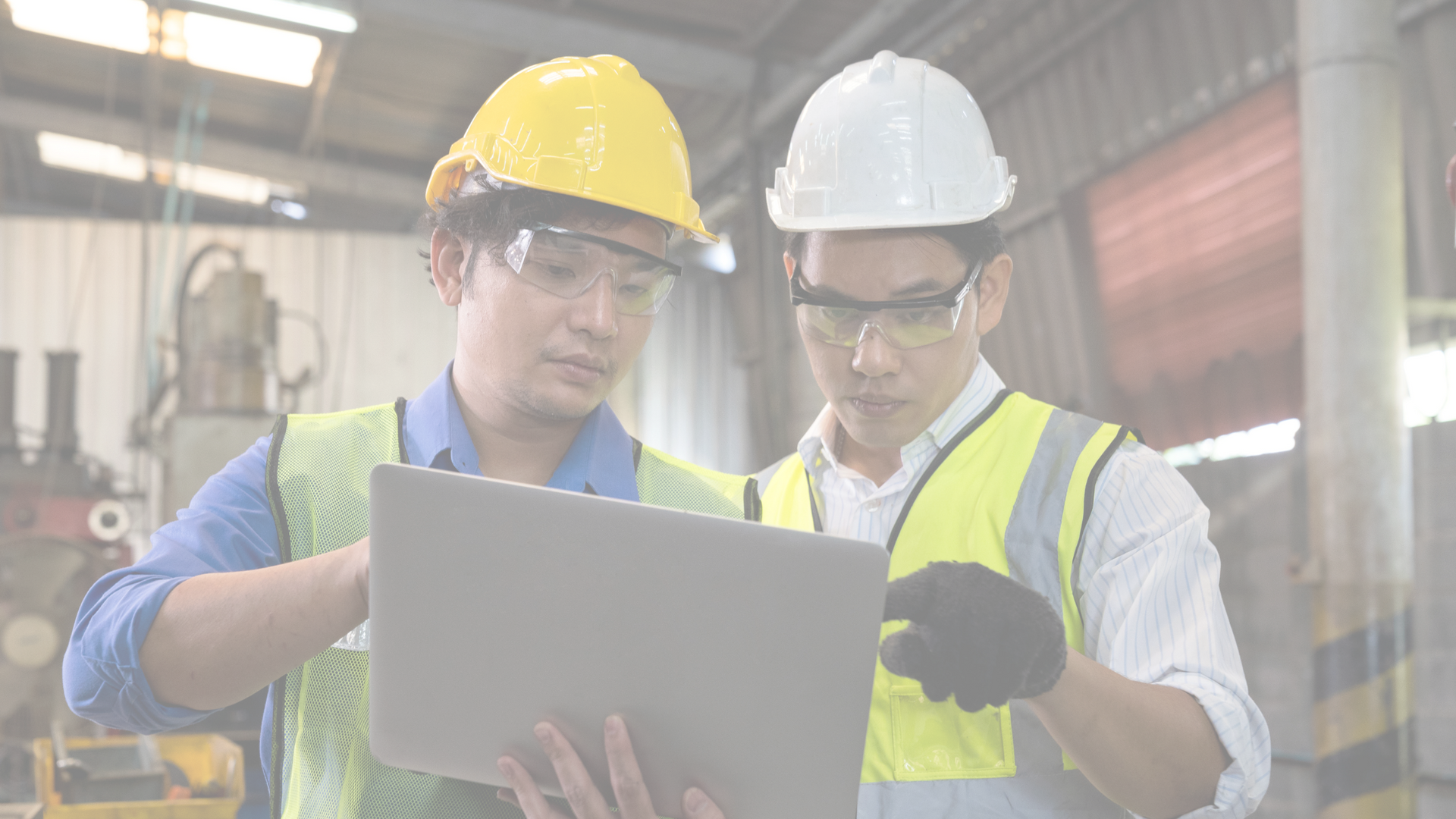 Two employees at work, wearing hard hats, safety glasses, and safety vests, study a laptop screen.