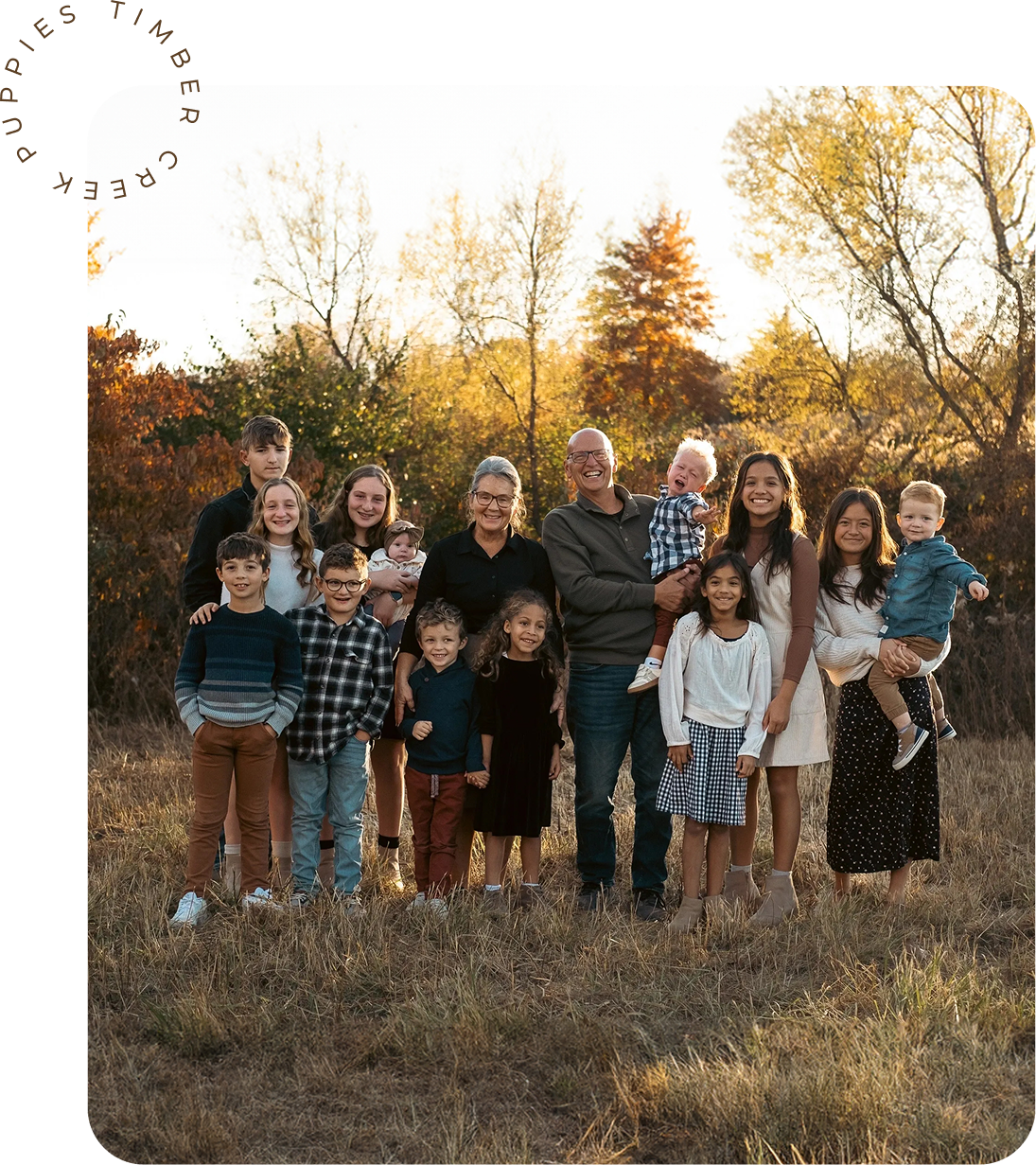 A large multi-generational family poses for a portrait in a sunlit, grassy field during the fall.
