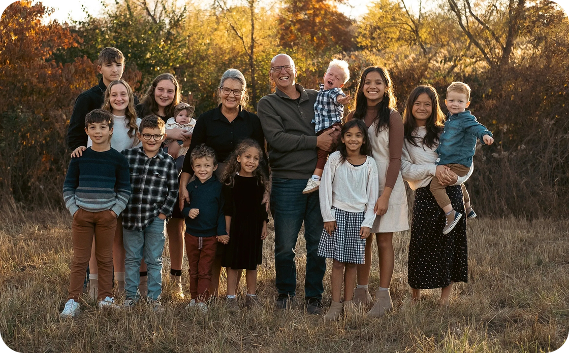 A large family posing for a group portrait in a field during golden hour with autumn trees in the background.