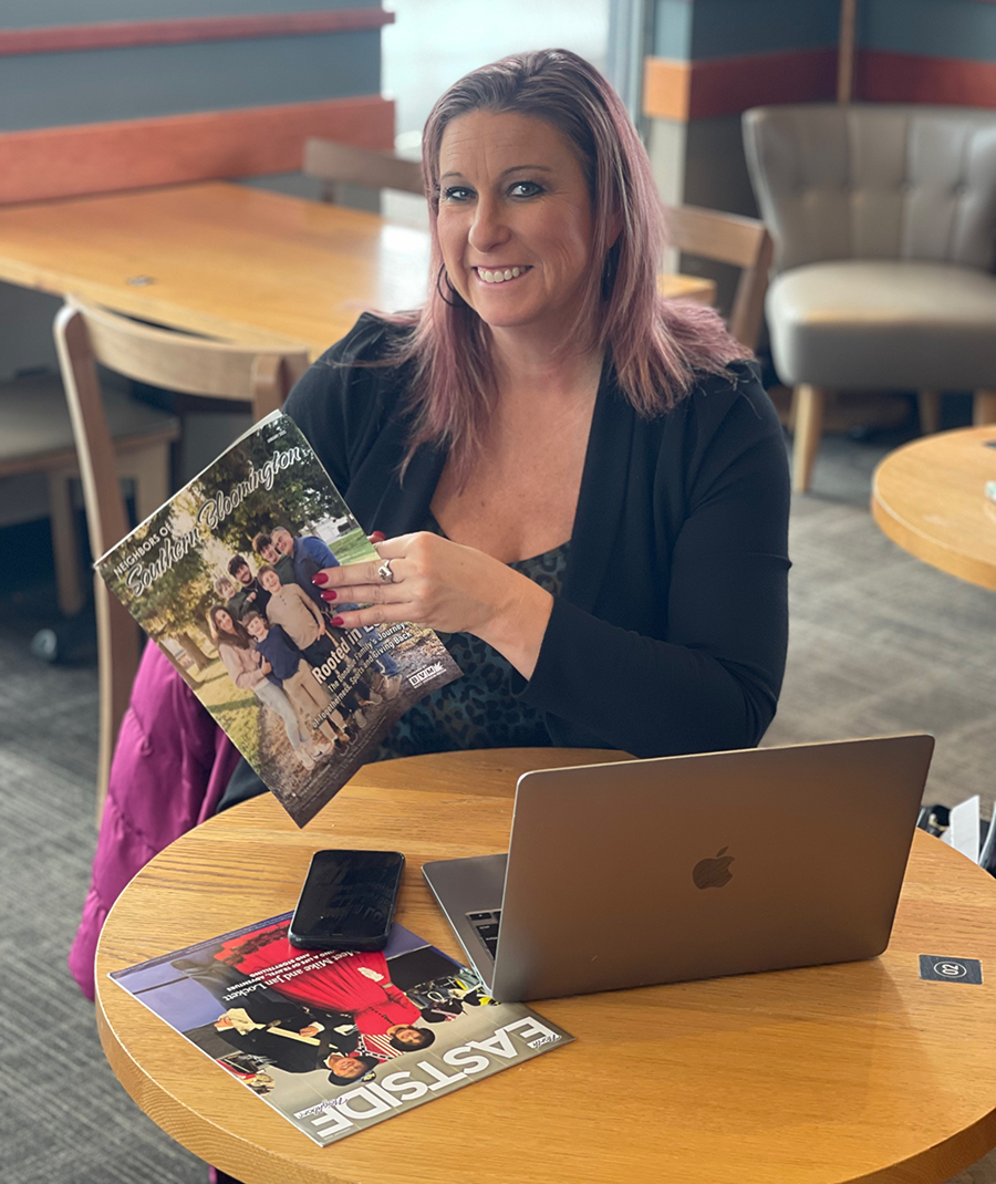 Woman smiling, holding a magazine at a cafe table with a laptop and phone.