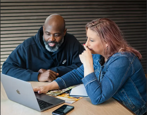 Two people look at a laptop. Woman points, man watches. They are in a room, light wood table and backdrop.