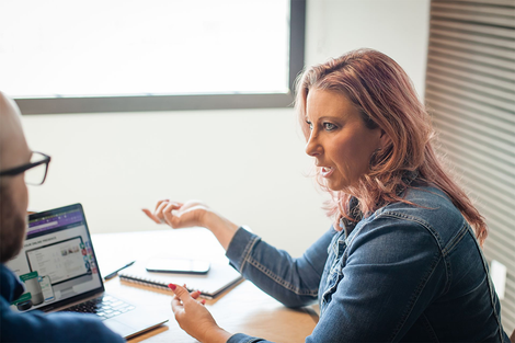 Woman with pink hair gesturing while speaking to a person at a laptop in a meeting room.