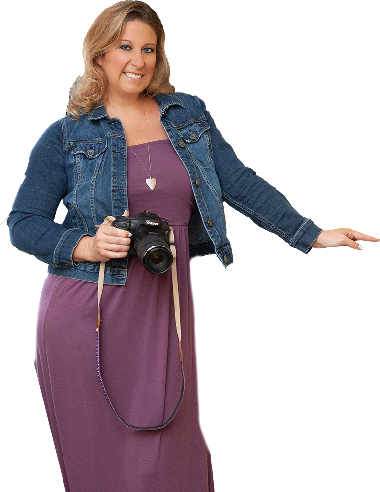 Woman in denim jacket and purple dress, holding a camera, smiling.