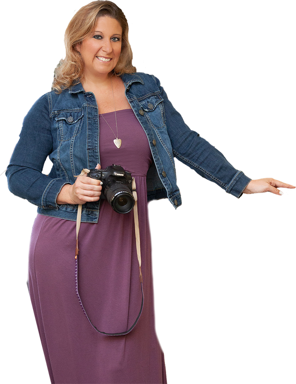 Woman in denim jacket and purple dress, holding a camera, smiling.