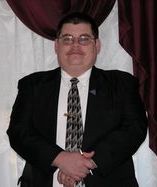 Two men in suits smile, standing by a framed portrait in a room with patterned wallpaper.