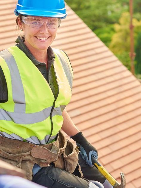 A woman wearing a hard hat and safety vest is sitting on a roof holding a hammer and smiling.