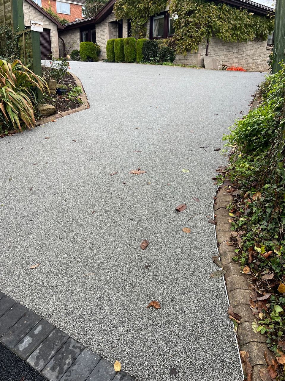 Grey resin driveway leading uphill towards a bungalow with lots of greenery around